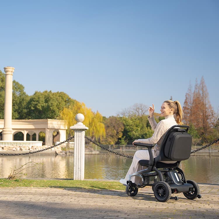 Revamed Robooter E80 Elektrische rolstoel zwarte bekleding en frame met verloop naar wit vooraan het frame. Voetensteun in het midden van de rolstoel en bestuuringsjoystick aan het uiteinde van de armleuning. Sfeerbeeld onderweg bij een park.